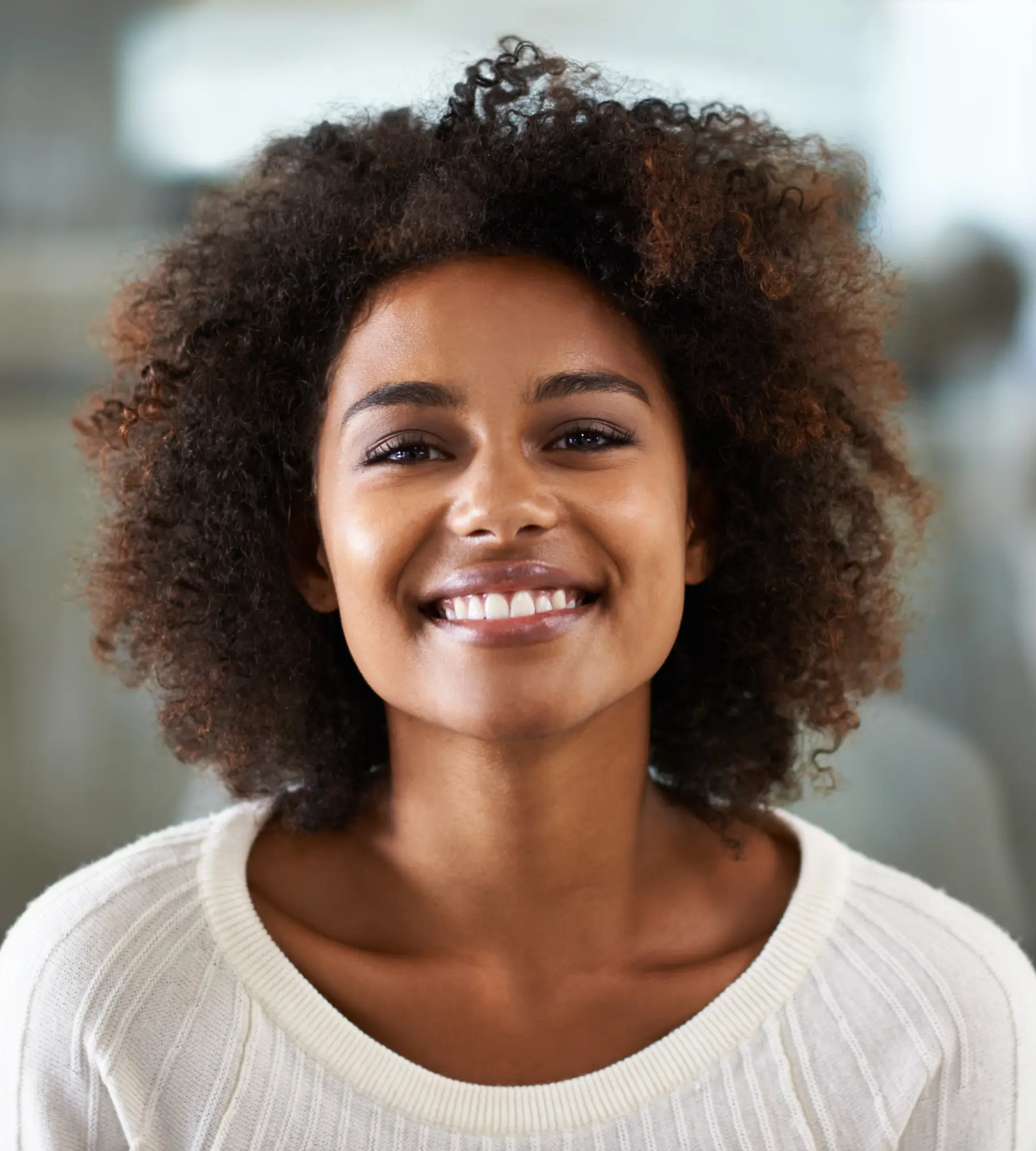 A person with curly hair smiles warmly at the camera, wearing a white sweater.