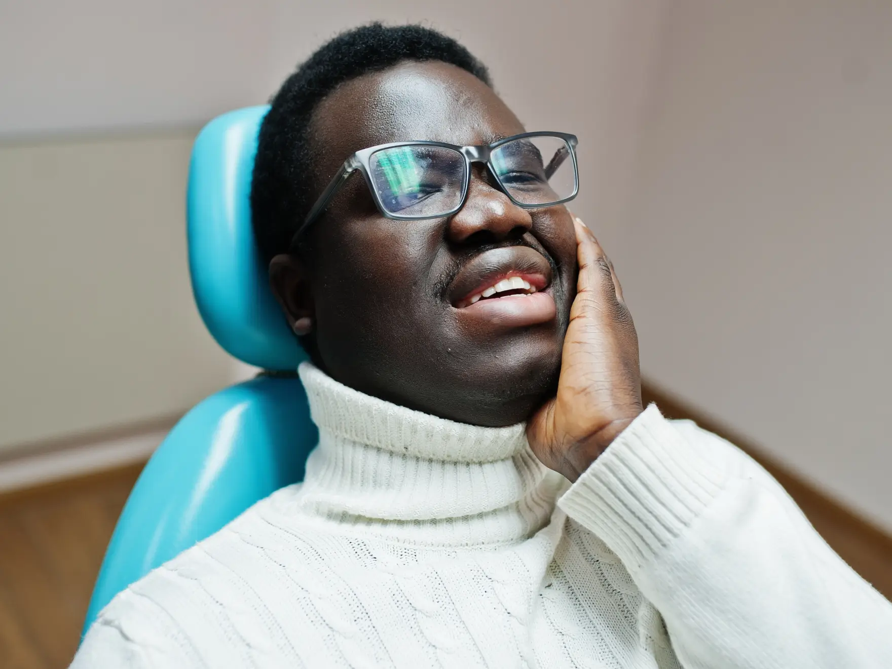 A man in glasses holds his cheek, sitting in a dental chair, appearing in pain.