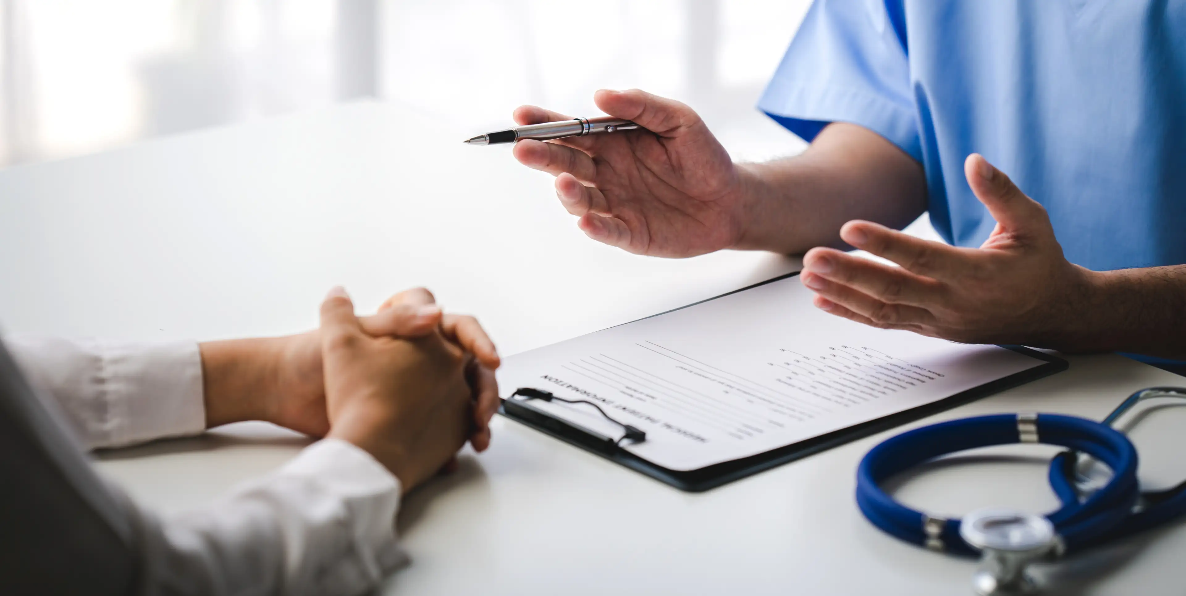 Doctor in blue scrubs explaining a form to a patient with clasped hands at a white table.