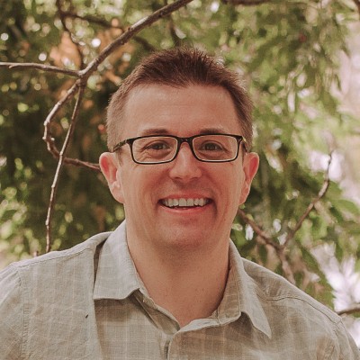 Smiling man wearing glasses and a light-colored shirt standing outdoors with trees in the background.