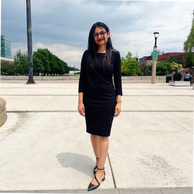 Woman with long black hair and glasses wearing a black dress and heels standing on a paved area with trees and buildings in the background.