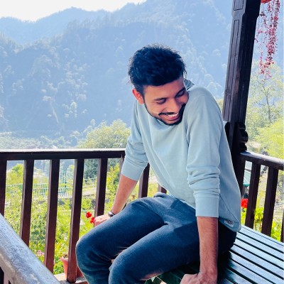 Smiling young man in a light blue sweater sitting on a bench on a balcony overlooking green hills.