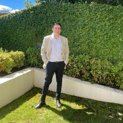 Young man wearing a beige blazer, white shirt, and black pants standing on grass in front of a lush green hedge under a partly cloudy sky.