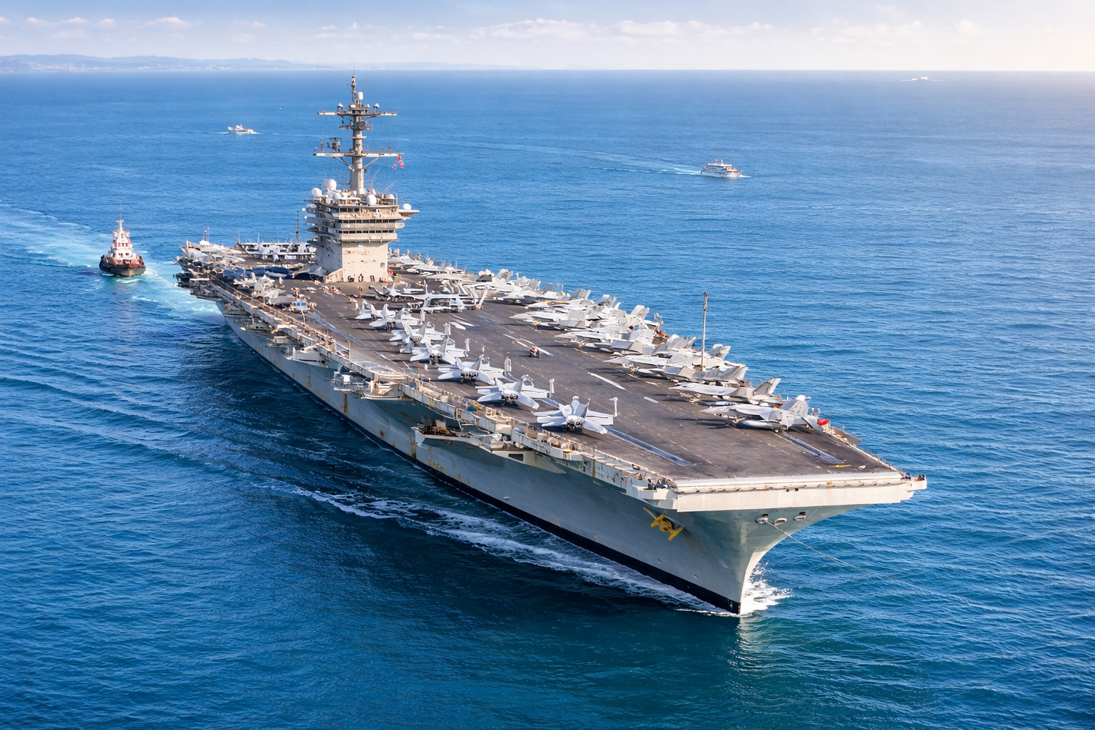 Aircraft carrier sailing in blue ocean with multiple fighter jets lined up on deck and a tugboat nearby.