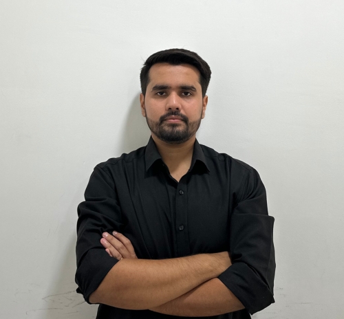 Man with short dark hair and beard wearing a black shirt with arms crossed against a white wall.
