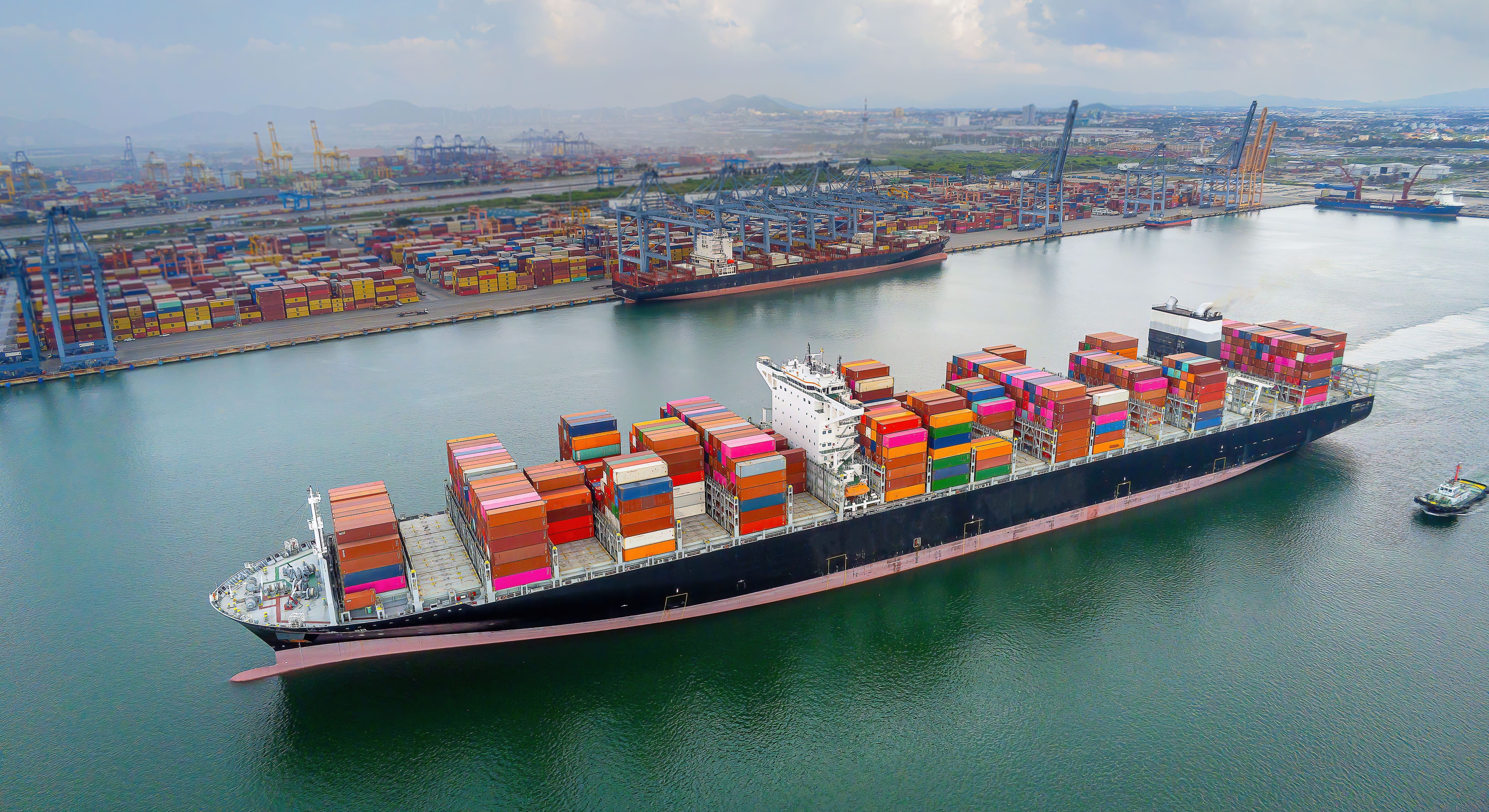 Large container ship loaded with colorful shipping containers departing a busy port with cranes and stacked containers.