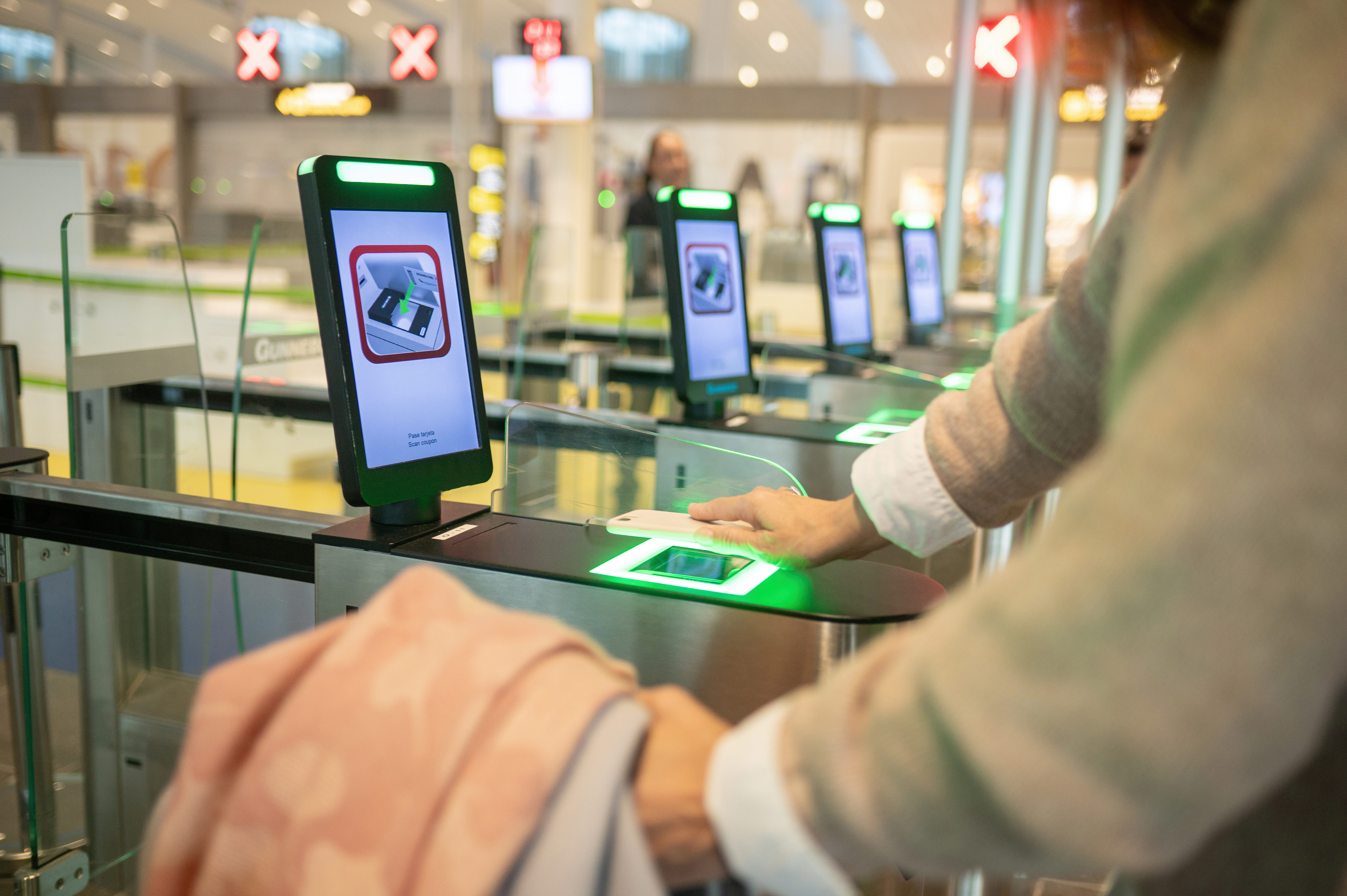 Person scanning a mobile phone at a green-lit self-check-in kiosk in an airport security area with red X signs in the background.