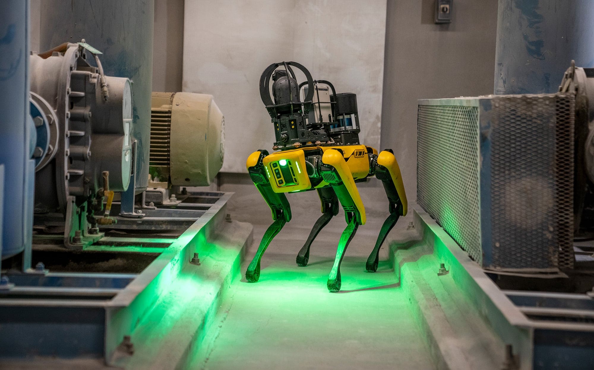 Yellow quadruped robot with sensors walking between industrial machinery with green lights on the floor.