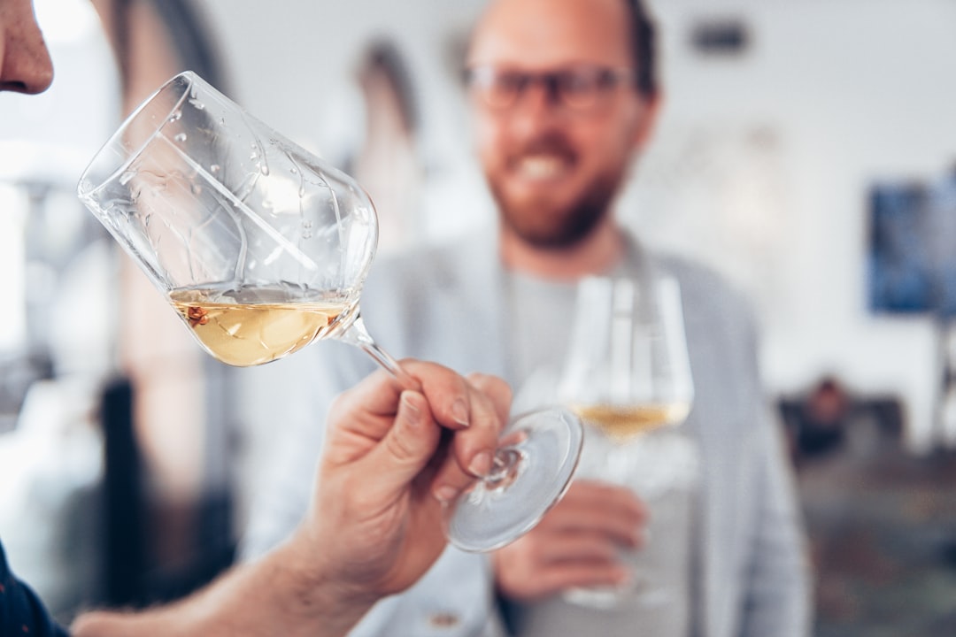 Das Bild zeigt zwei Männer, die an einer Weinverkostung teilnehmen. Im Vordergrund hält eine Person ein Weinglas mit weißem Wein und betrachtet es aufmerksam, während die andere Person im Hintergrund lächelnd ebenfalls ein Weinglas in der Hand hält. Beide tragen legere Kleidung und befinden sich in einem hellen, modernen Raum mit unscharfem Hintergrund, der auf eine gesellige und entspannte Atmosphäre hinweist. Dieses Bild passt perfekt zum Abschnitt über die Weinverkostung im Blog-Beitrag und illustriert die gehobene und lehrreiche Natur einer solchen Veranstaltung. Es unterstreicht die Aspekte der Geselligkeit, des Lernens und des Genusses, die bei einer Weinverkostung im Vordergrund stehen. Solche Events bieten den Mitarbeitern die Möglichkeit, sich in einer entspannten Umgebung zu vernetzen und ihre sensorischen Fähigkeiten zu schärfen.