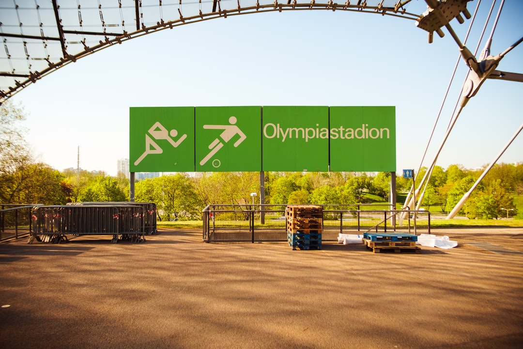 Das Bild zeigt das Olympiastadion mit einem großen, grünen Schild im Vordergrund, auf dem Piktogramme für verschiedene Sportarten sowie der Schriftzug Olympiastadion zu sehen sind. Das Stadion ist von einer offenen Struktur mit Stahlträgern umgeben, und im Hintergrund erstreckt sich eine grüne Parklandschaft unter einem klaren blauen Himmel. Einige Paletten und Absperrungen stehen auf dem Asphaltboden vor dem Schild. Dieses Bild passt perfekt zum Abschnitt über die Firmenolympiade im Blog-Beitrag und illustriert die Idee eines sportlichen Wettbewerbs in einer inspirierenden Umgebung. Es unterstreicht die Aspekte des freundlichen Wettbewerbs, der Teamarbeit und des Gemeinschaftsgefühls, die durch eine Firmenolympiade gefördert werden. Solche Veranstaltungen bieten eine abwechslungsreiche Möglichkeit, Mitarbeiter zu motivieren und gleichzeitig Spaß und körperliche Aktivität zu fördern.