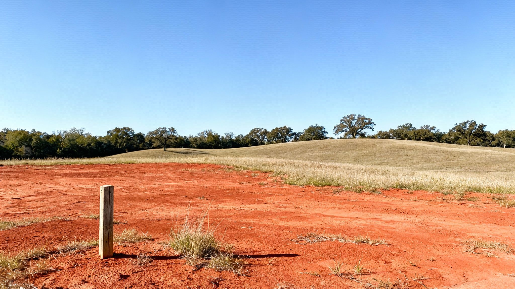 A landscape view featuring red dirt in the foreground, grassy hills, and a line of trees under a clear blue sky.