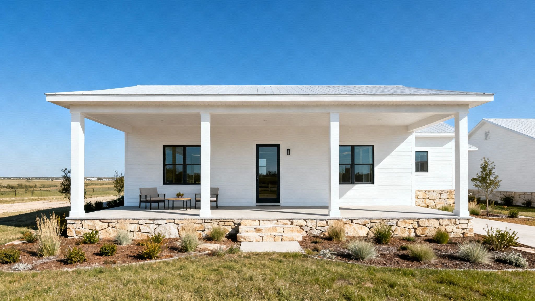 A modern white farmhouse with a covered porch, black windows, stone base, and green landscaping.