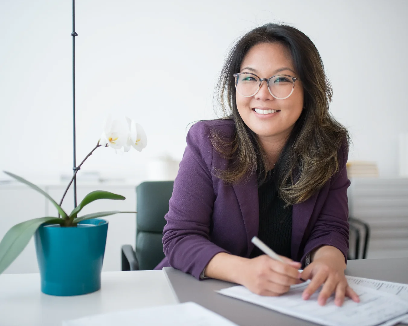 Professional woman in glasses and a purple blazer writing at a desk beside an orchid.
