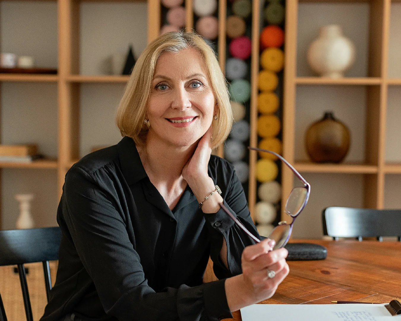 Professional woman seated at a table, holding eyeglasses in a warm office setting.