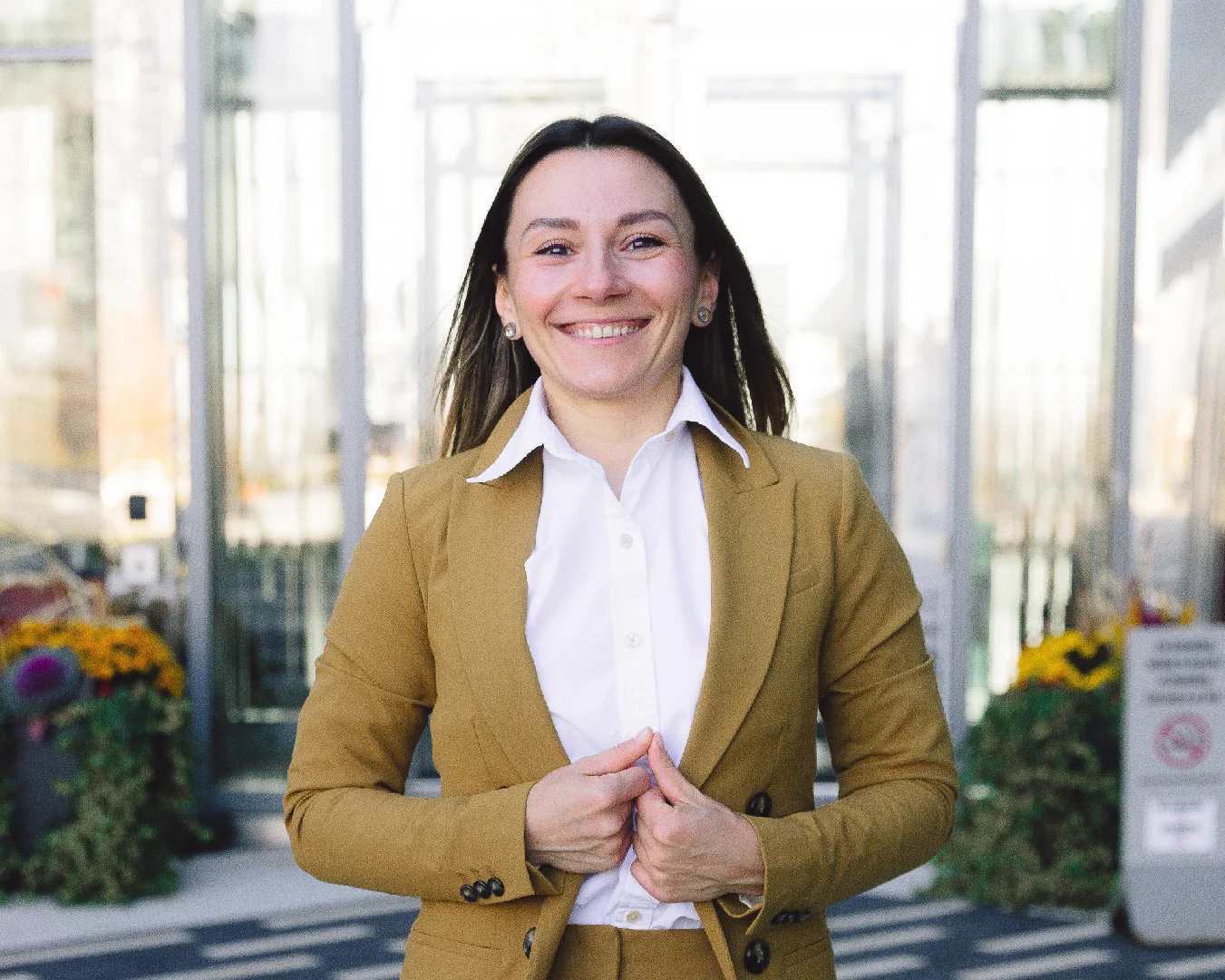 Professional woman smiling in a tan suit near a bright glass building entrance.