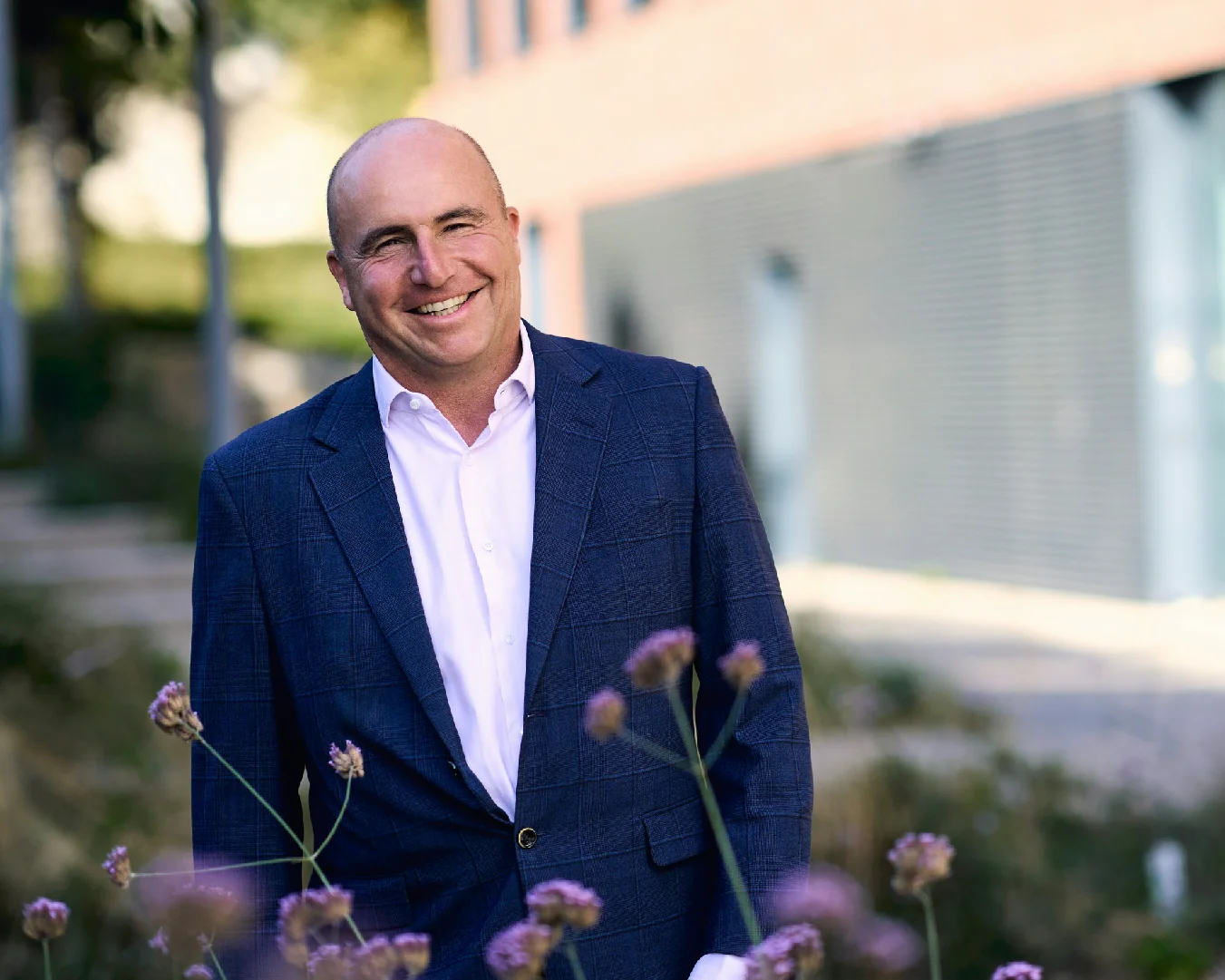 Professional man in a blue suit smiling outdoors in a landscaped setting.