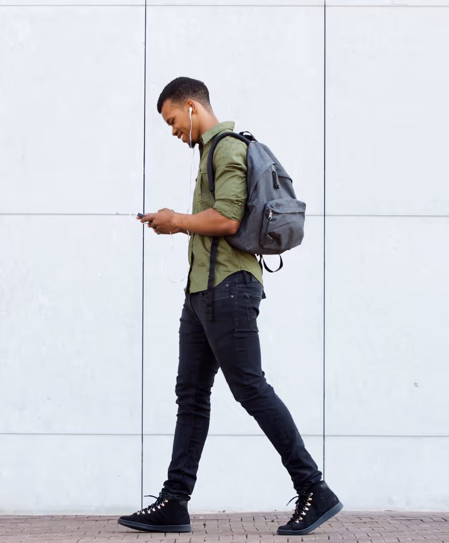 Young man walking outdoors with a gray backpack, wearing earphones and looking at his smartphone.
