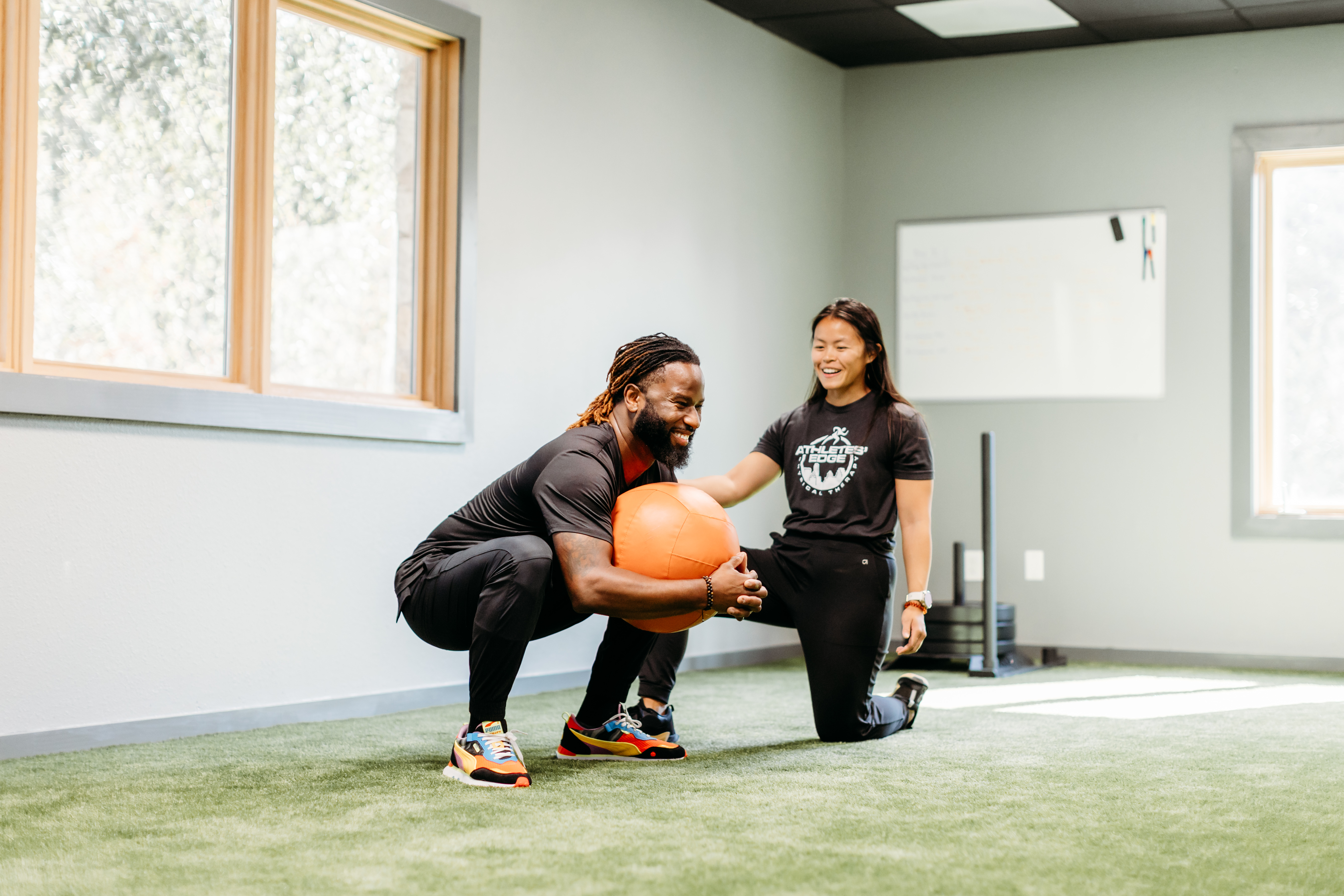 Trainer in black points to correct deadlift form while man in light blue shirt and white cap lifts barbell in gym.