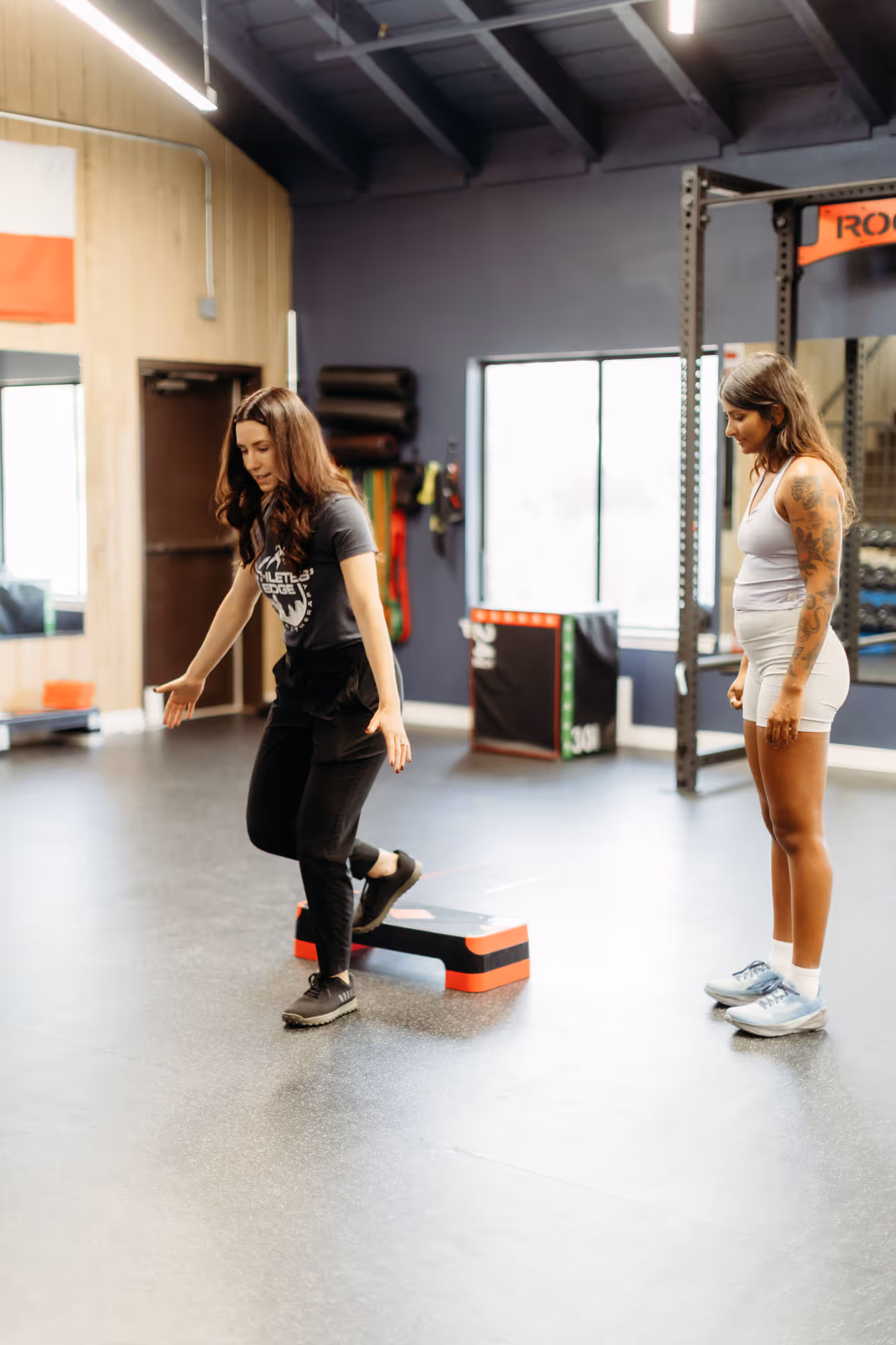 Trainer guides woman stepping onto aerobic step in bright gym with fitness equipment.