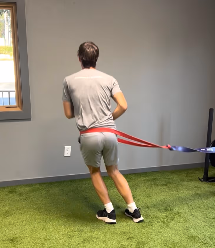 Man in gray shirt and shorts doing resistance band lateral walk exercise on green turf indoors.