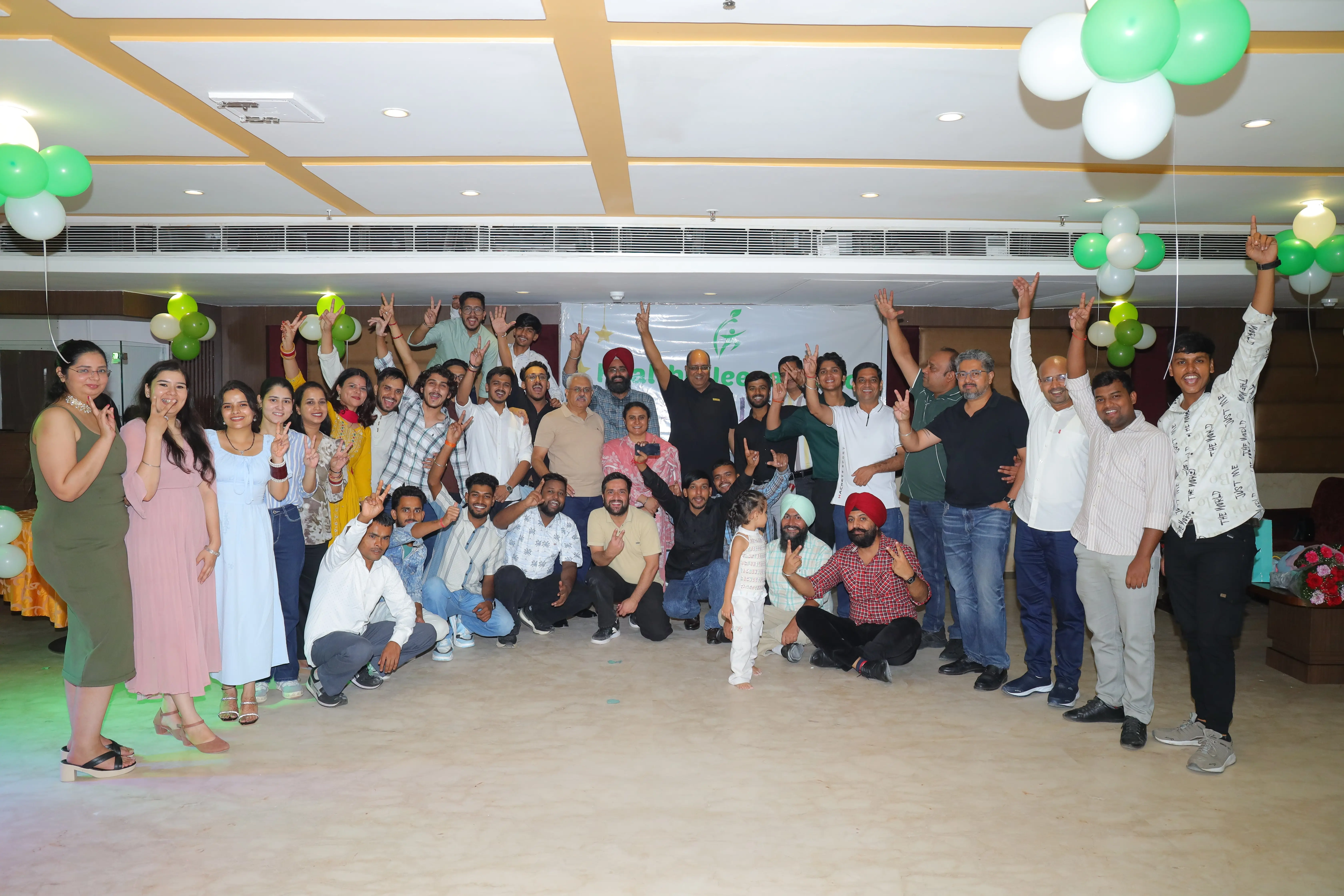 A large group of Healthy Jeena Sikho team members posing together indoors during a celebration event. The room is decorated with green and white balloons, and everyone is smiling, making peace signs, or raising their hands in excitement. The group includes men, women, and a child, standing and kneeling in front of a white banner with the Healthy Jeena Sikho logo.