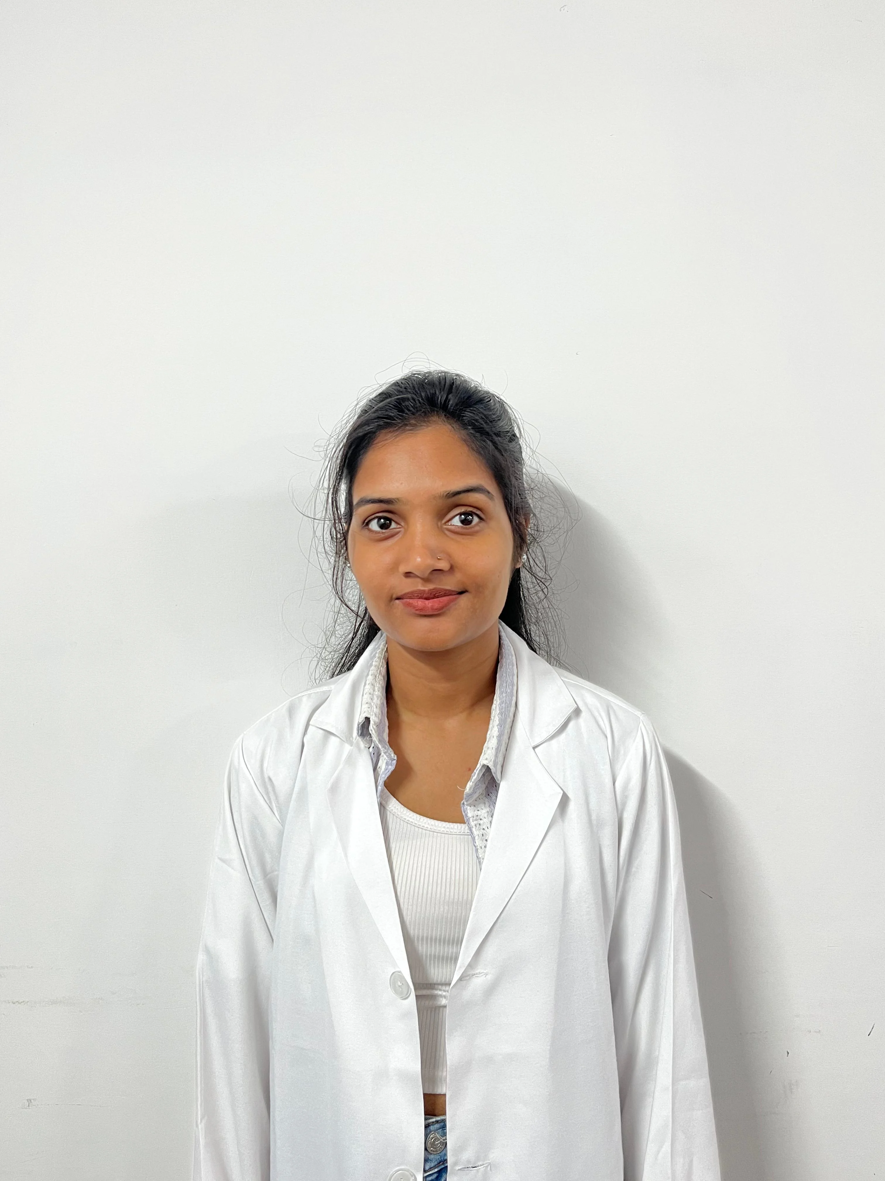 A medium, eye-level shot of a young woman with dark hair pulled back, wearing a white lab coat over an olive-green t-shirt. She is standing against a plain white or light grey wall. She is looking directly at the camera with a neutral expression.