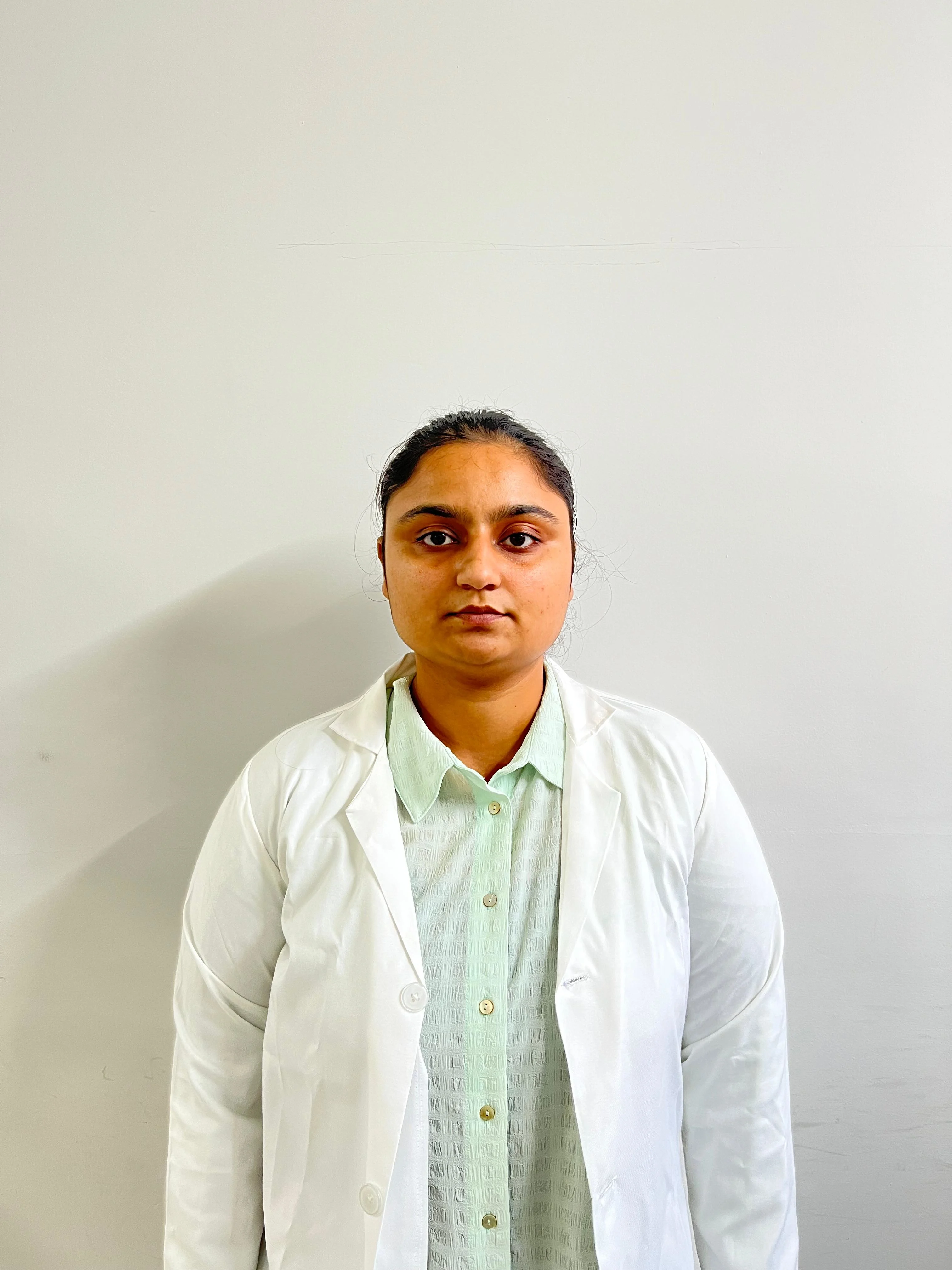 A medium, eye-level shot of a young woman with dark hair pulled back, wearing a white lab coat over an olive-green t-shirt. She is standing against a plain white or light grey wall. She is looking directly at the camera with a neutral expression.