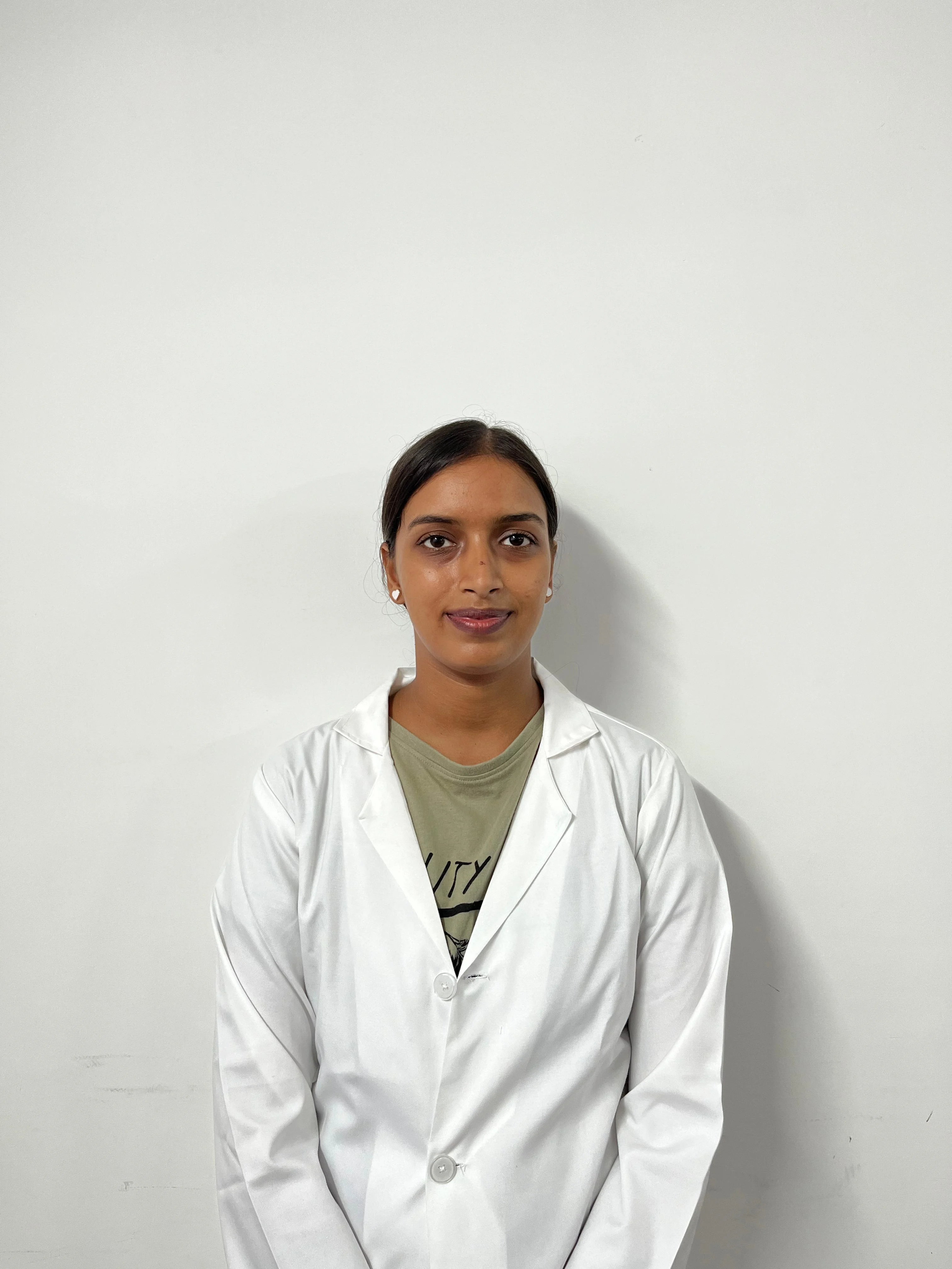 A medium, eye-level shot of a young woman with dark hair pulled back, wearing a white lab coat over an olive-green t-shirt. She is standing against a plain white or light grey wall. She is looking directly at the camera with a neutral expression.