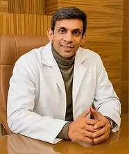 A smiling male doctor in a white lab coat and a turtleneck, sitting at a desk with hands clasped.