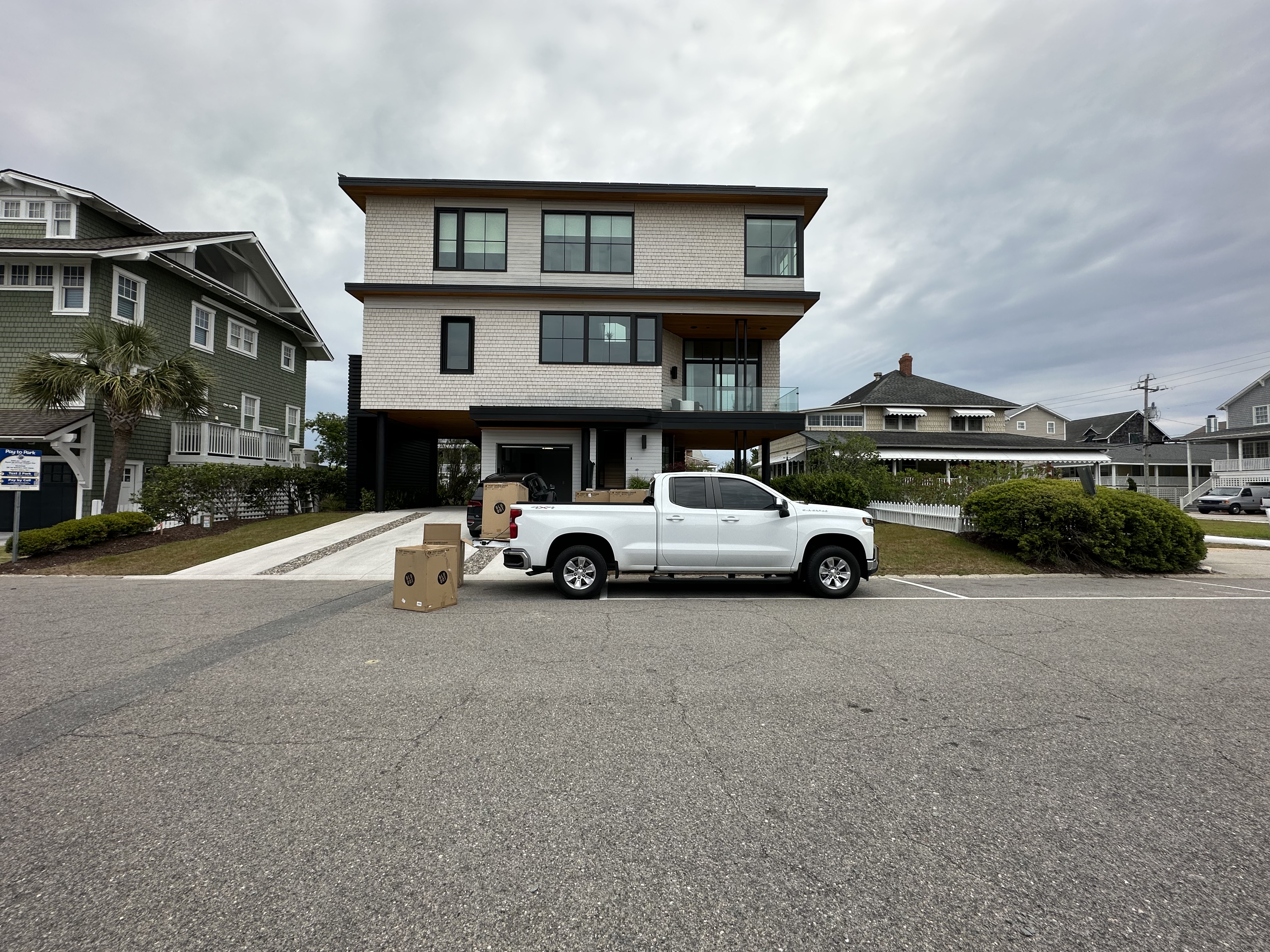 Modern three-story house with white exterior and large windows, a white pickup truck parked in front, and cardboard boxes on the street near the truck.