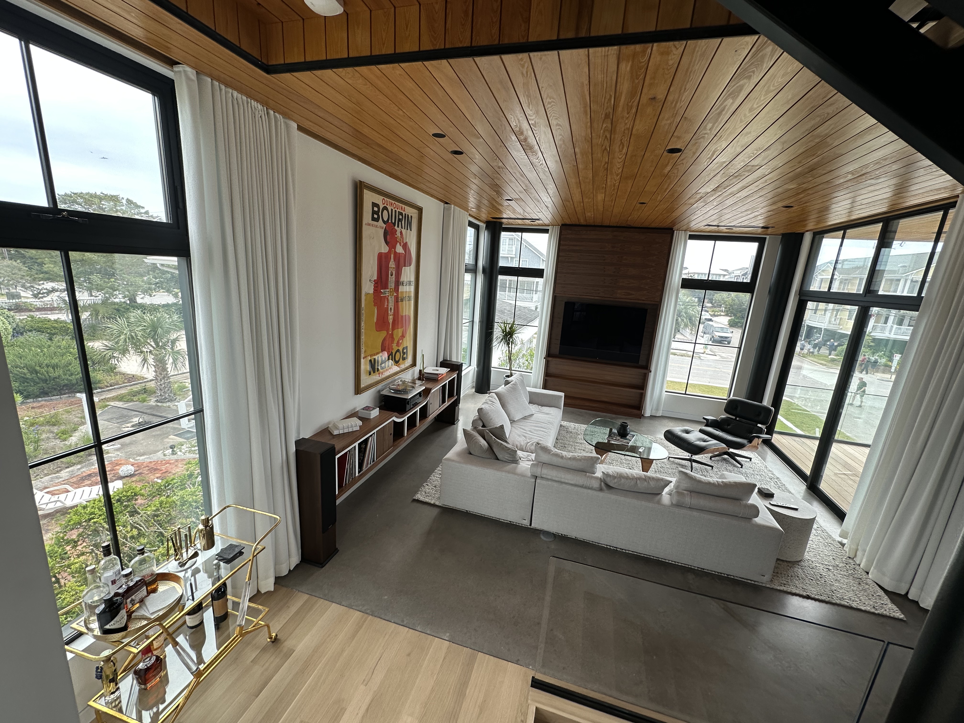 Modern living room with large windows, white sectional sofa, black lounge chair, glass coffee table, wooden ceiling, and a gold bar cart.