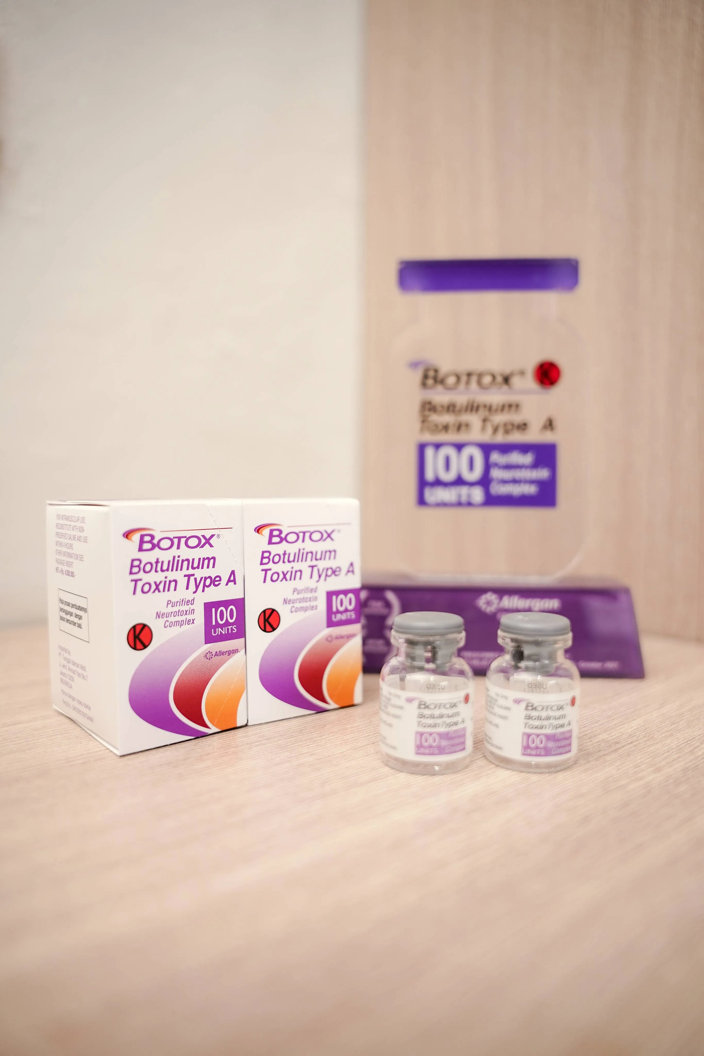 Two boxes and two vials of Botox Botulinum Toxin Type A, 100 units, on a wooden surface with a blurred background display.