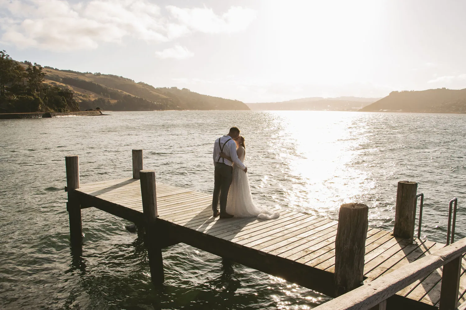 Couple embracing on wooden pier extending into calm water with rolling hills meeting the shoreline under bright sky with soft clouds