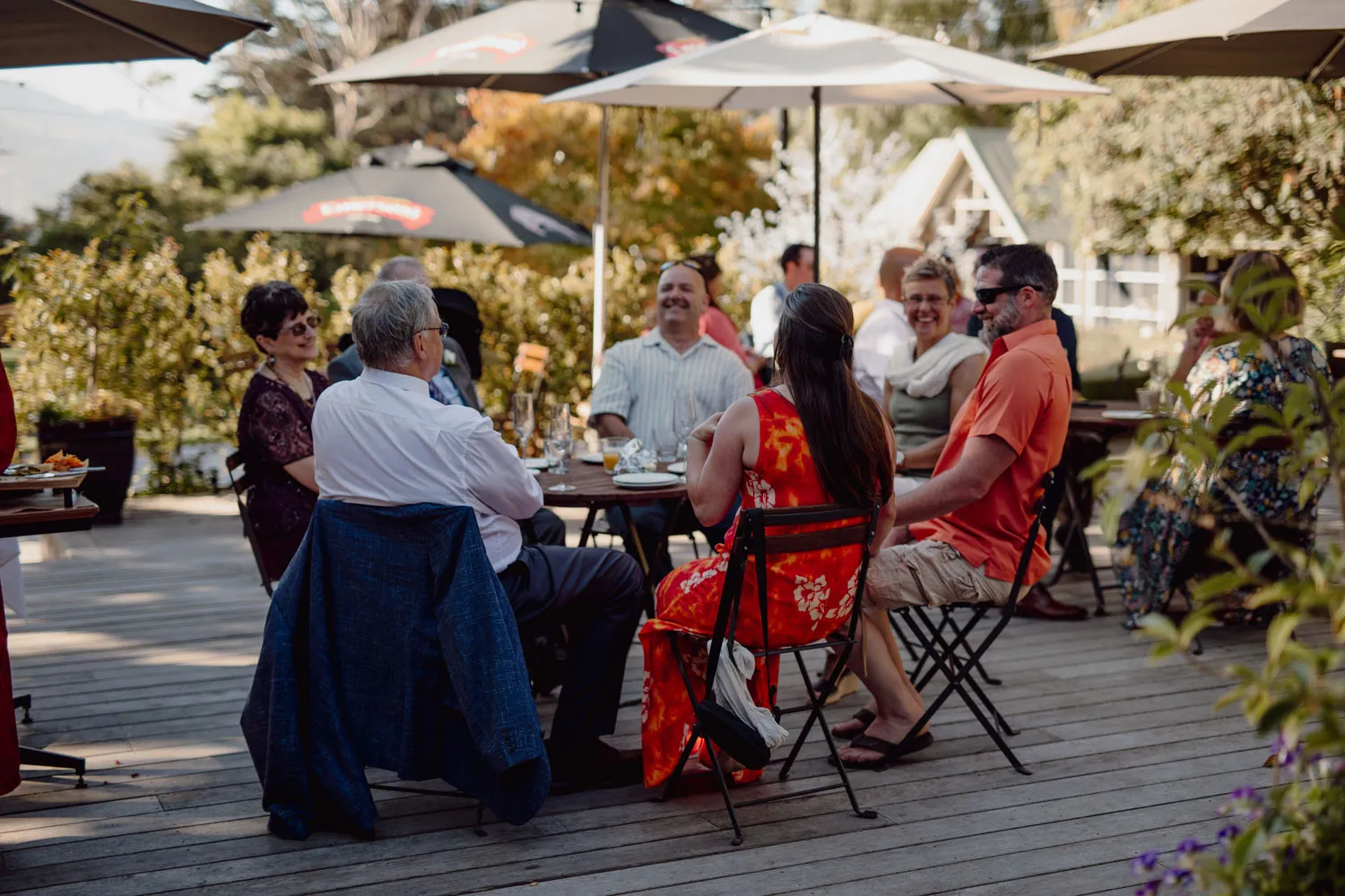 Group of people seated at tables on wooden deck for outdoor event, surrounded by lush greenery with A-frame building in background