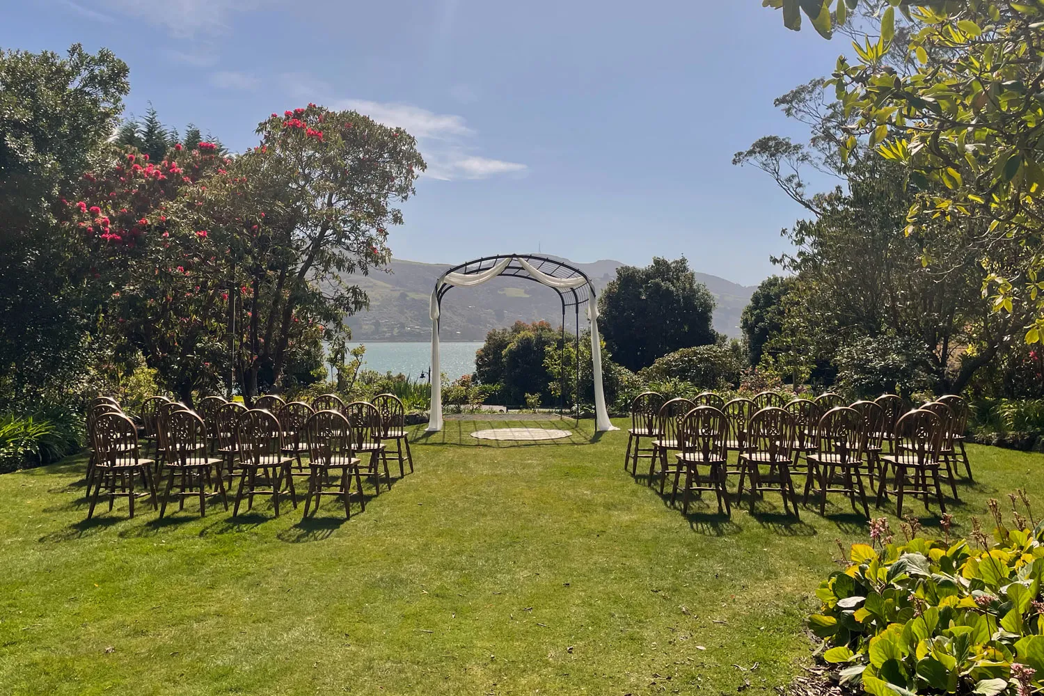 Outdoor wedding ceremony setup with white chairs on green lawn facing black metal arch draped with white fabric, overlooking calm water and green hills at Glenfalloch Gardens in Dunedin.