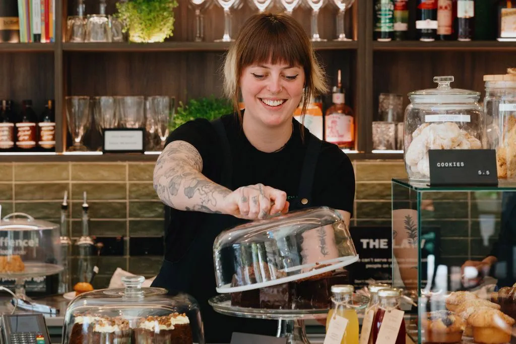 Smiling female barista lifting a glass cover off a chocolate cake in a cozy café with pastries and drinks displayed.