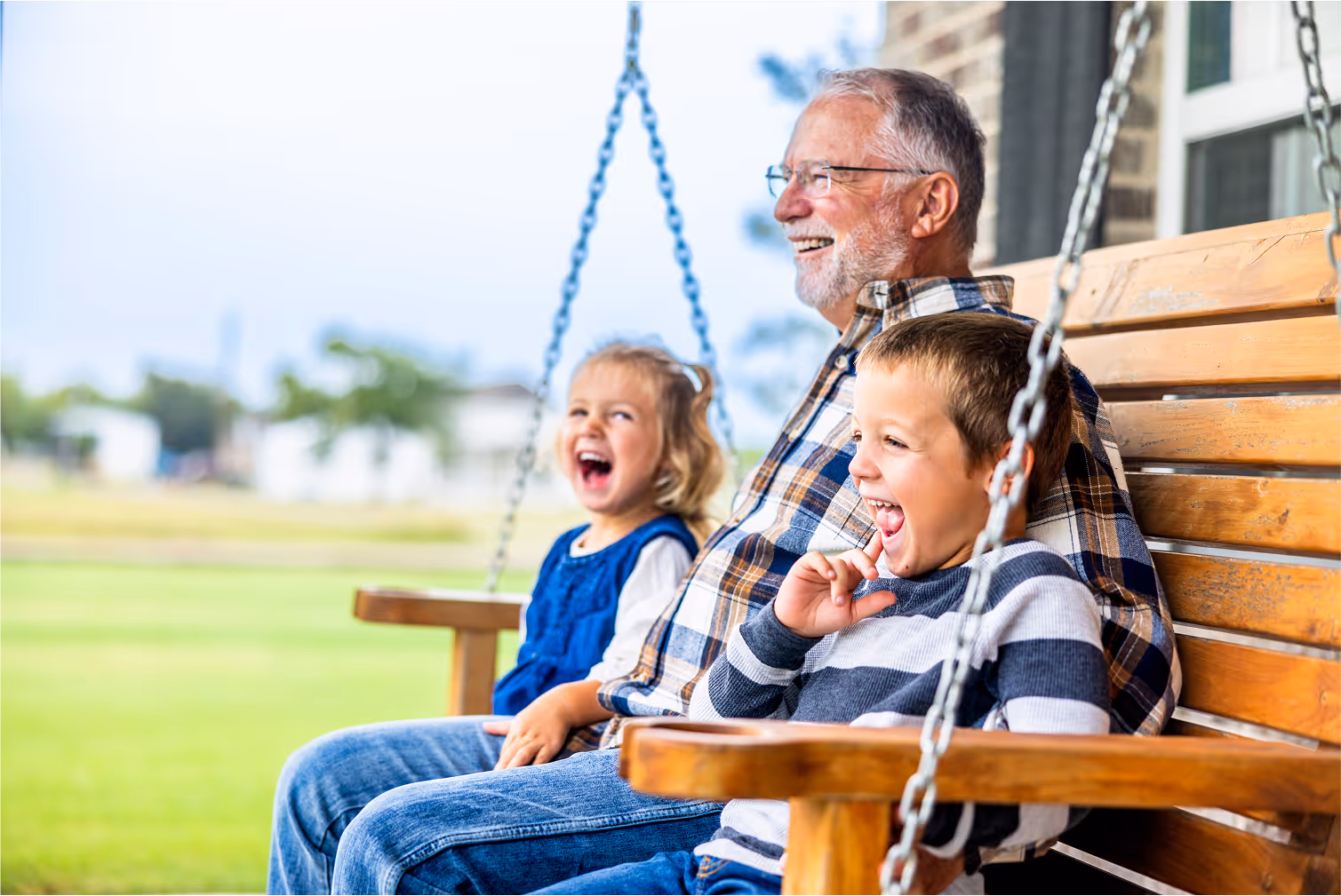 Older man smiling while sitting on a porch swing with two laughing young children on a bright day.