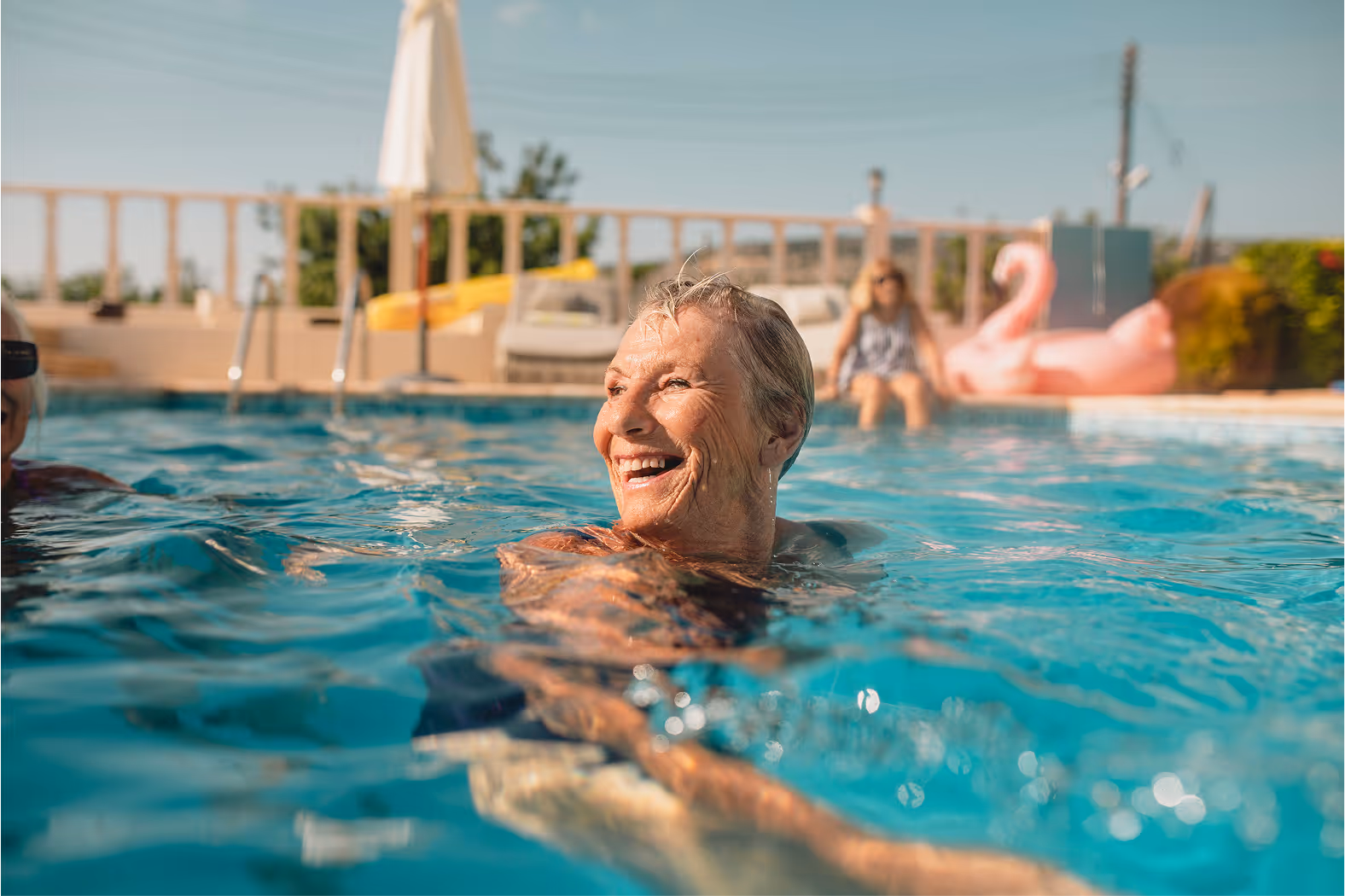 Smiling older person swimming in an outdoor pool on a sunny day, with others relaxing in the background.