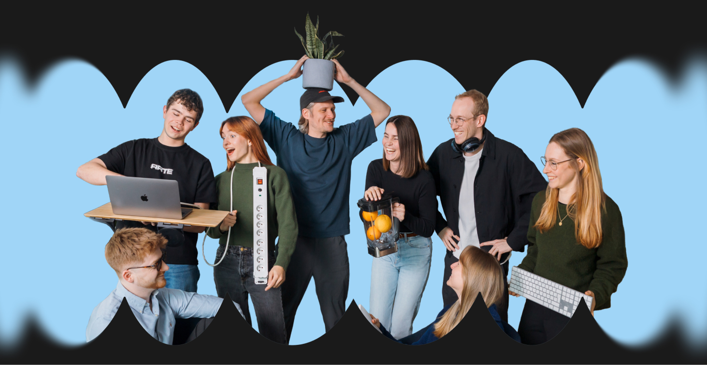 Group of seven young adults smiling and interacting, holding various items including laptop, power strip, potted plant, blender with oranges, and keyboard against a blue background.