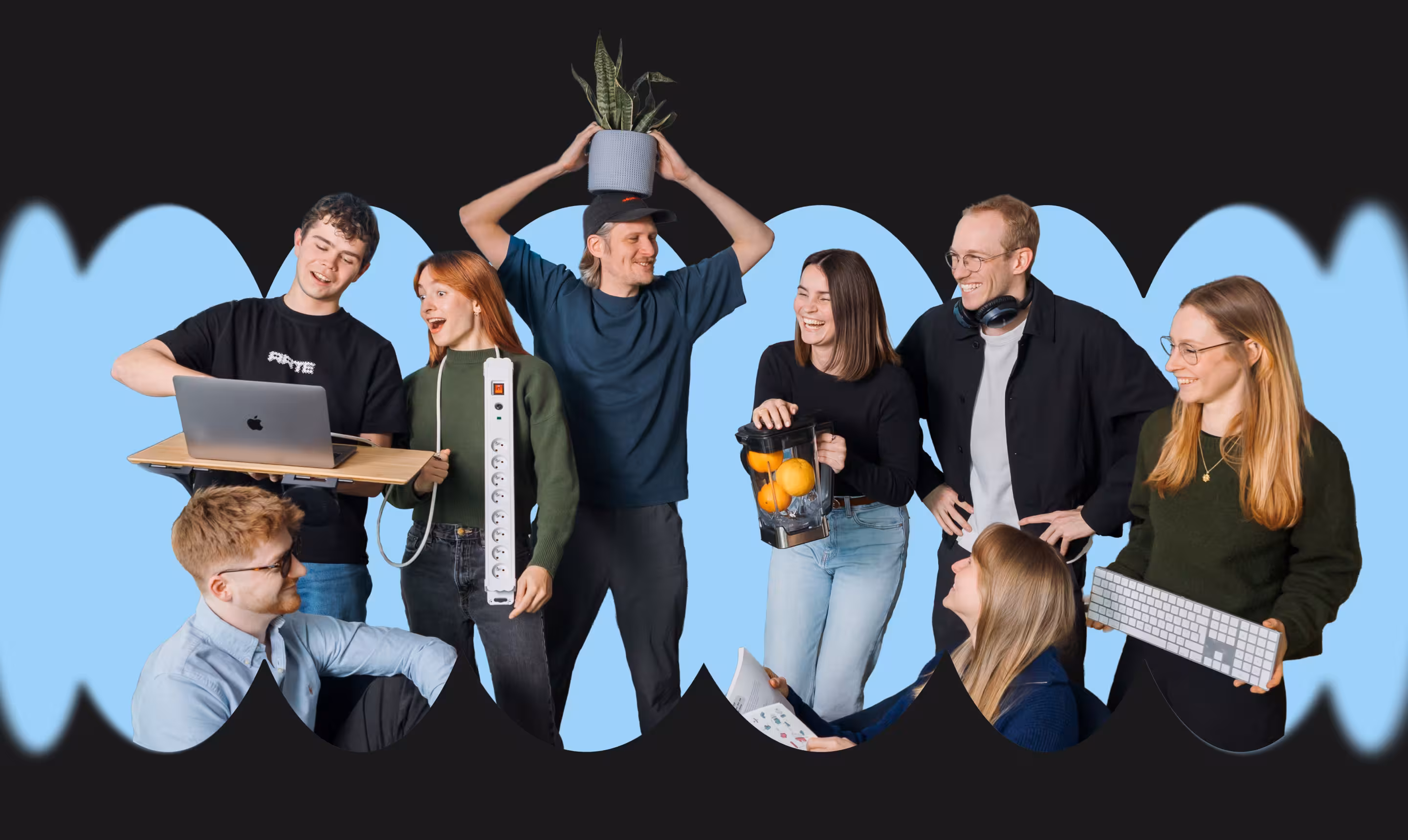 Group of seven young adults smiling and interacting, holding various items including laptop, power strip, potted plant, blender with oranges, and keyboard against a blue background.