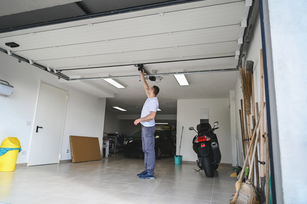A man in a white shirt opens a garage door, revealing a car and a motorcycle inside.