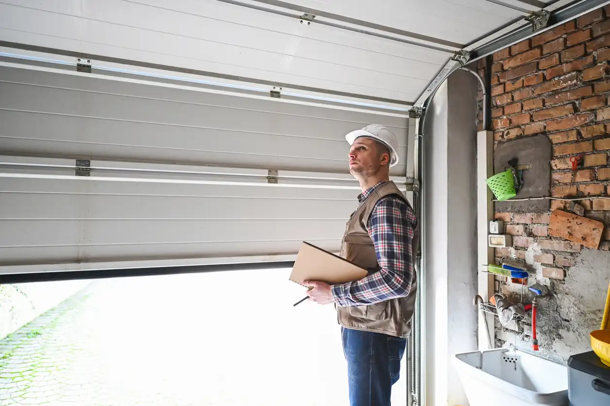 Person in hard hat inspecting garage door mechanism inside brick-walled garage with tools and sink