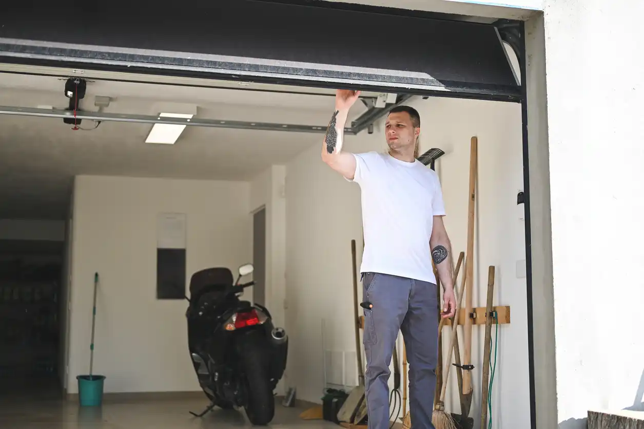 A man in a white t-shirt stands inside a garage with a motorbike and tools, reaching up to the garage door.