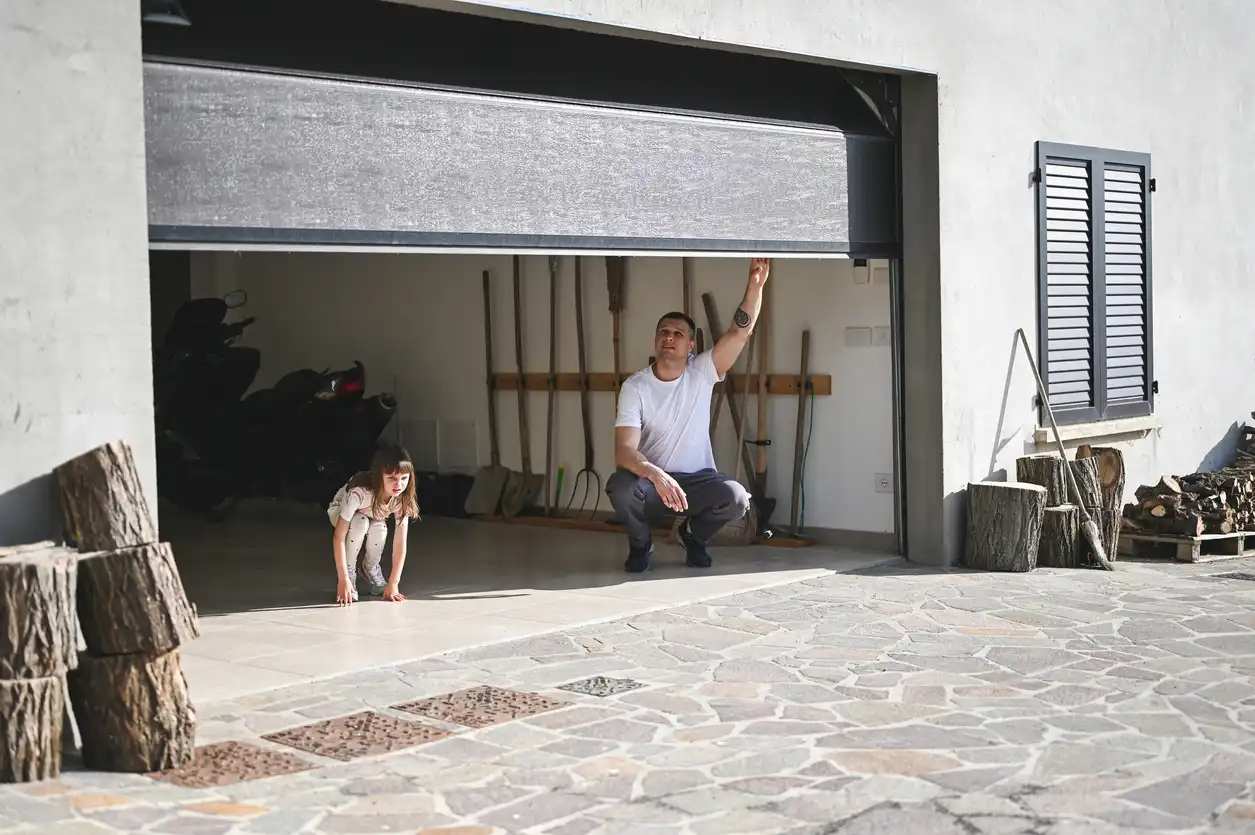A man and a young child are in a garage with the garage door partially open. The man, wearing a white t-shirt and dark pants, is in a squatting position, reaching up towards the garage door. The child, wearing a light-colored dress, is also squatting further back in the garage. Inside the garage, there are tools leaning against a wall and what appears to be a motorcycle. Outside the garage, there are stacks of cut logs and a window with dark shutters on the right side of the building. The ground in front of the garage is paved with light-colored stones.