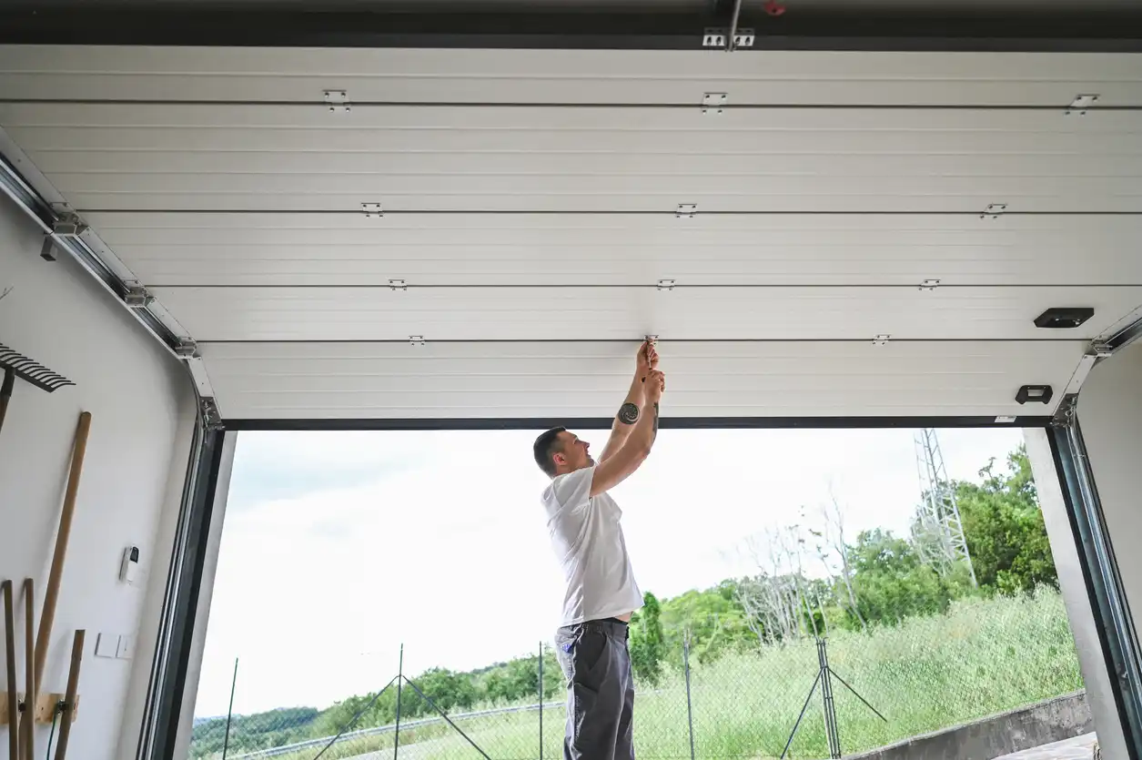 A man in a white t-shirt reaches up to adjust the ceiling mechanism of a white garage door from inside.