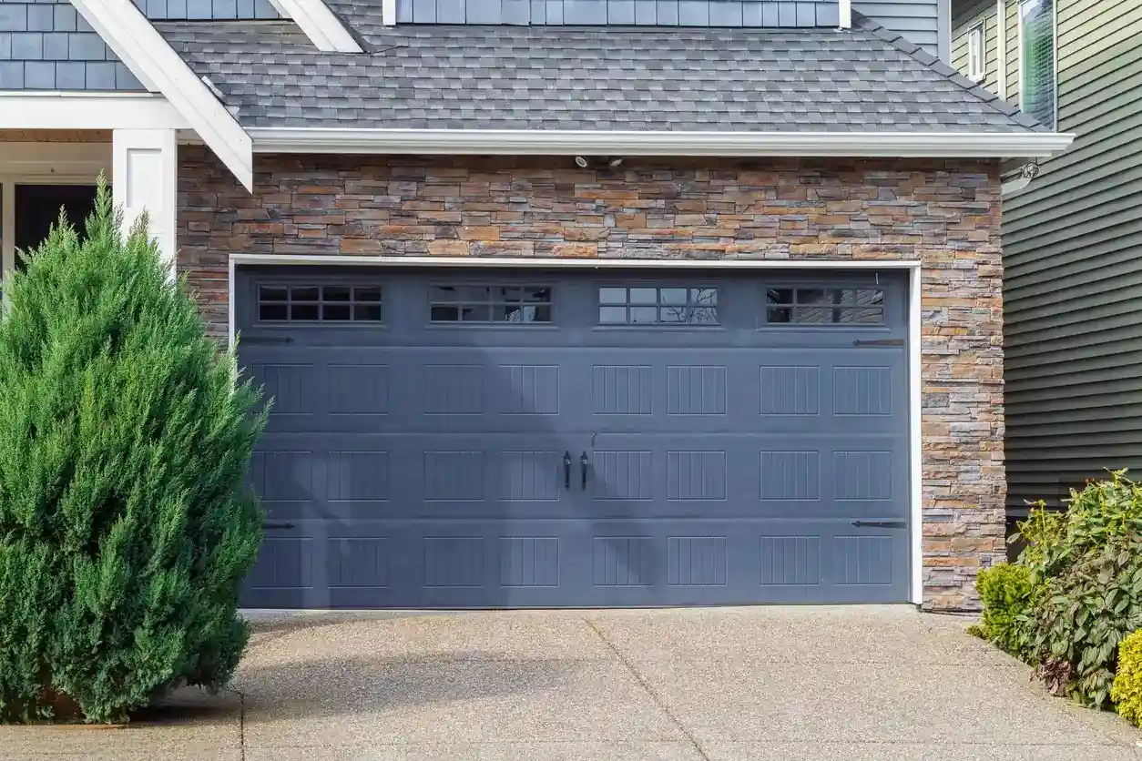 A grey two-car garage door with windows, set in a house with stone and siding.