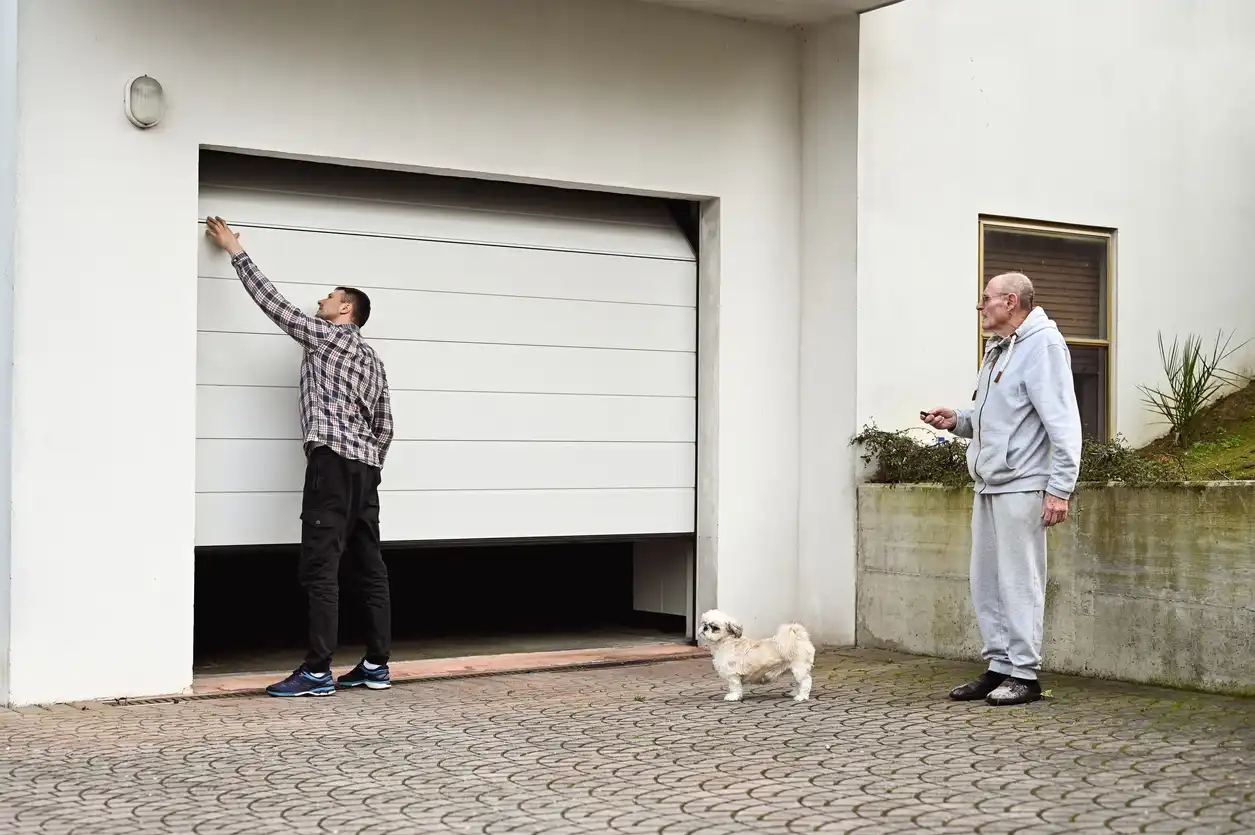 A man adjusts a white garage door as it opens, with an older man and a small dog nearby.