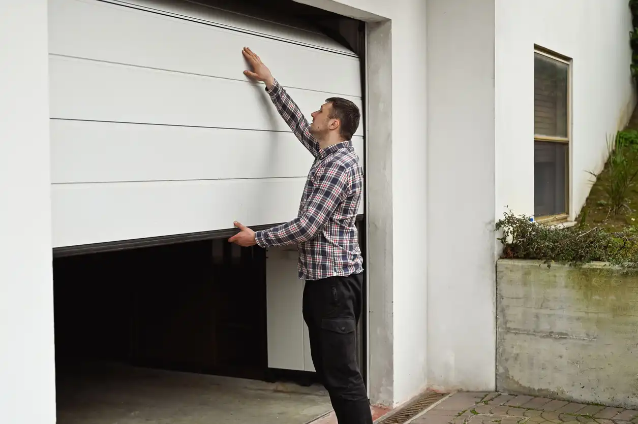 A man in a plaid shirt adjusts a white garage door as it opens.