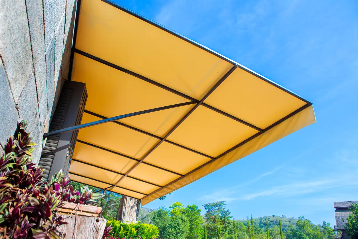 Yellow awning shading garden with plants under clear blue sky, trees and hills in background