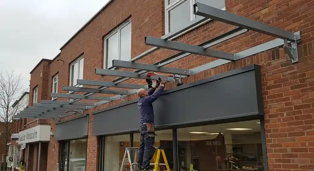 A worker on a ladder installs a metal awning frame above a storefront, attaching a crossbeam to the main structure.